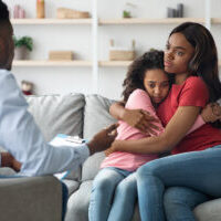 Black woman hugging her scared daughter while attending child psychologist, african american mother and child teenager having therapy session with male psychotherapist. PTSD in children
