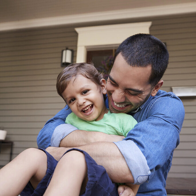Father Hugging Son As They Sit On Porch Of House Together